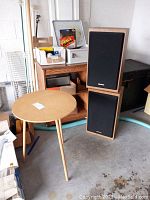 Wooden shelf, round wooden three-legged table, two Kenwood speakers stacked vertically in a garage or storage space