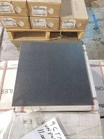 Close-up of single square porcelain tile in matte black finish on top of stacked cartons with labels visible indicating tile size and quantity