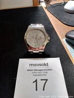 Front view of a silver toned wristwatch showing the white dial with three subdials and Arabic numeral 12, sitting on a black mat with auction lot card number 17.