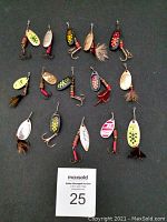 Top-down view of 14 vintage Mepps spinner fishing lures arranged in rows on black surface, showing variety of colors, spinner blade shapes, feathered treble hooks, and signs of use.