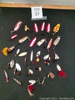 Top-down view of 28 assorted vintage fishing lures arranged on a black background showing varied sizes, shapes, and colors including red, white, silver, gold, and yellow. Some lures have feather attachments on hooks.
