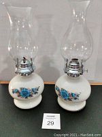 Pair of vintage oil lamps with white ceramic bases featuring blue floral patterns, silver metal burner assemblies, and clear glass chimneys.