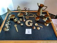 Box of assorted used brass items displayed on black mat, including figurines, pitcher, bowls, key-shaped items, candle holders and ladle.