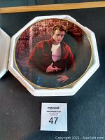 Front of octagonal James Dean collector plate with image of James Dean in a red jacket and white shirt, gold rimmed, displayed in a white box.
