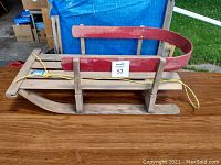 Wooden sleigh with curved red backrest, wooden runners, placed on wooden table outdoors.