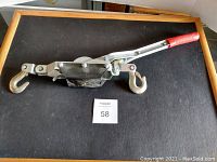 Photo of the opposite side of come along hand winch with red handle on black surface showing hooks and central mechanism.