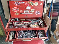 Front view of vintage red metal tool chest with stickers and top drawer open showing hand drills, tape measures, and small tools