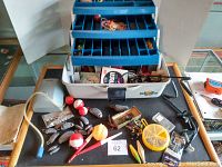 Photo showing open fishing tackle box with all accessories in and around the box including reels, bobbers, lines, clips, weights, and lures.