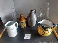 Photo showing six bottles and pitchers including four glass bottles, one brown ceramic jug with cork, one yellow decorative porcelain pitcher, and one white porcelain pitcher with flower motif on a black surface.