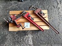 Photo showing one vintage red bench vise and two red vintage pipe wrenches on a wooden pallet placed on concrete floor.