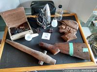 Full overview of lot items on black table: vintage travel iron, wooden carvings including a rifle and club, wool bust, glass bottle, frosted glass shade, plug adapters, and accessory box.