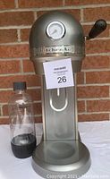 Front and side view of KitchenAid Effervescent soda maker and included plastic bottle on white cloth, set against brick wall.