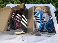 Two Adidas rugby cleats boxes open side by side, containing a black/red pair and a blue/white pair of cleats placed on a white surface outdoors.