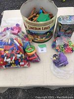 Full lot of creative toys showing Lincoln Logs tin, bag of Lego bricks, Light Up Links, colorful rings, and small toys spread on white table.