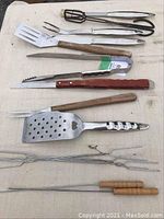 Set of eight BBQ utensils arranged on a cloth surface including spatula, fork, tongs, and skewers with wooden and metal handles.