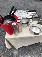 Photo of a table showing a red container holding metal pans and kitchen sieves, two square glass spice shakers with metal lids, and a small square metal mesh container.