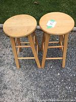 Photo showing two light-colored wooden stools with round seats, four legs, and cross bars, placed outdoors on gravel ground.
