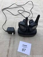 Two black Midland X-Tra Talk walkie talkies in charging base with attached power adapter on a light-colored fabric surface.