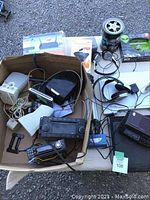 Top-down view of a cardboard box containing various electronic devices such as modems, routers, car stereos, wireless headphones, speakers, scanner, and cables.
