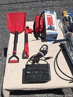 Full view of the lot on a beige surface on gravel showing the battery charger, three air pumps, snow shovel, and back saver attachment.