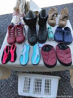 Top view of various boots and shoes arranged on a flat surface, including men's black steel toe boots, women's white cold weather boots, tan men's shoes, red athletic shoes, water shoes, slippers and blue slip-ons.