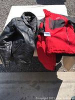 Men's black leather jacket and red and black winter jacket laid out side by side on white background outdoors.