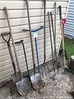 Full view of all tools lined up against wall including shovels, forks, rakes, and weed removers.