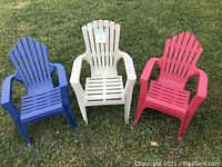 Three youth-sized Muskoka chairs in blue, white, and red placed on grass outdoors.