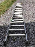 Full view of aluminum extension ladder laid flat on gravel surface, showing two 10 ft sections connected with sliding mechanism.