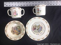Overview of four Bunnykins christening-themed china items: two double-handled cups, a shallow bowl, and a small plate with decorative borders and detailed colorful illustrations in Beatrix Potter style.
