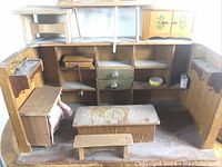 Wooden dollhouse furnishings arranged inside a wooden dollhouse structure showing shelves, drawers, a table, and a bench.