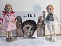 Two porcelain dolls flanking a hardcover book titled 'THE DOLL' showing a close-up of a doll's face on the cover.