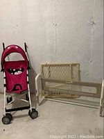 Cosco pink stroller with black and white striped handles and wheels, placed beside Safety 1st bed rail and wooden baby gate against a white wall.