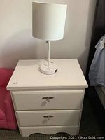 White nightstand with two drawers, crystal-style handles, and a white table lamp on top