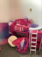 Shows a pink and purple doll bed, a white fluffy chair, a pink polka-dotted baby carrier, and a pink fabric doll tent or canopy folded on the floor