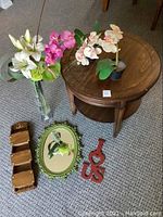 Overview of entire lot showing wood end table, glass vase with flowers, two artificial orchids, green oval mirror, red decorative piece, and stacked wooden trays.