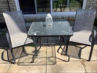 Outdoor patio set with 34x34 square glass top table and two chairs placed on a concrete patio, showing the set in full with sunlight casting shadows.