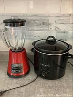 Black Crock-Pot slow cooker next to red Oster blender on a countertop.