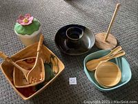 Photo of assorted serving ware including ceramic bowls with vegetable designs, wooden salad utensils, ceramic cupcake container, black candle holder with wooden base and spindle, turquoise serving dish with bamboo bowls and spatulas.