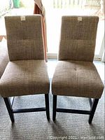 Front view of two identical bar height chairs with upholstered brown fabric seats and backs, standing side by side indoors.