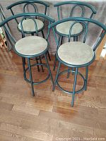 Four metal swivel bar stools with light beige microfiber round seats and black metal frames, arranged on wooden floor.