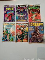 Front view of 6 comic books laid flat showing their colorful vintage illustrated covers including Man-Bat #1, Stalker #1 and #4, Justice League #12, Sgt. Rock #287, and Creepy #145.