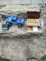 Blue vintage Radio Shack 1987 Malibu 4x4 off roader RC truck with wired black remote controller and wooden battery charger box with lid open, displayed on a stone surface outdoors.