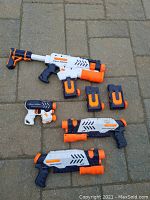 Photo showing four Supersoaker water guns and two belt clip loaders placed on outdoor pavement surface.