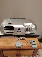 Silver Epson Powerlite S1+ projector placed on a wooden table with Philips DVD player underneath. White Epson remote and gray AV adapter also on the table.