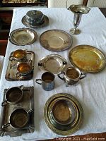 Full view of all the assorted silverplate items including trays, cups, creamer, sugar bowl, bowl, and goblet arranged on a white cloth.