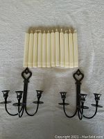 Two black metal wall-mounted triple candle holders with heart-shaped decorative top and 12 cream-colored smokeless taper candles laid flat on a white textured surface.