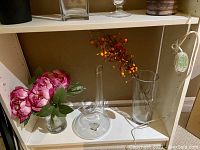 Shelf with artificial pink flowers in small glass vase, clear glass decanter style vase, clear tall cylindrical vase with faux berry stem, and clear glass tall cylinder vase.