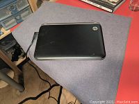 Black HP Mini laptop closed on a gray surface, connected to power cable, showing top cover with HP logo and some wear marks.