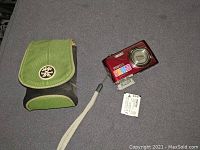 Front and side view of red Pentax Optio P70 camera with lens extended, green and black fabric carrying case, and battery.